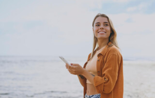 A woman walking on the beach and using travel apps on her phone.