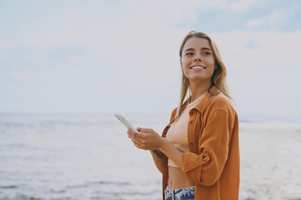 A woman walking on the beach and using travel apps on her phone.